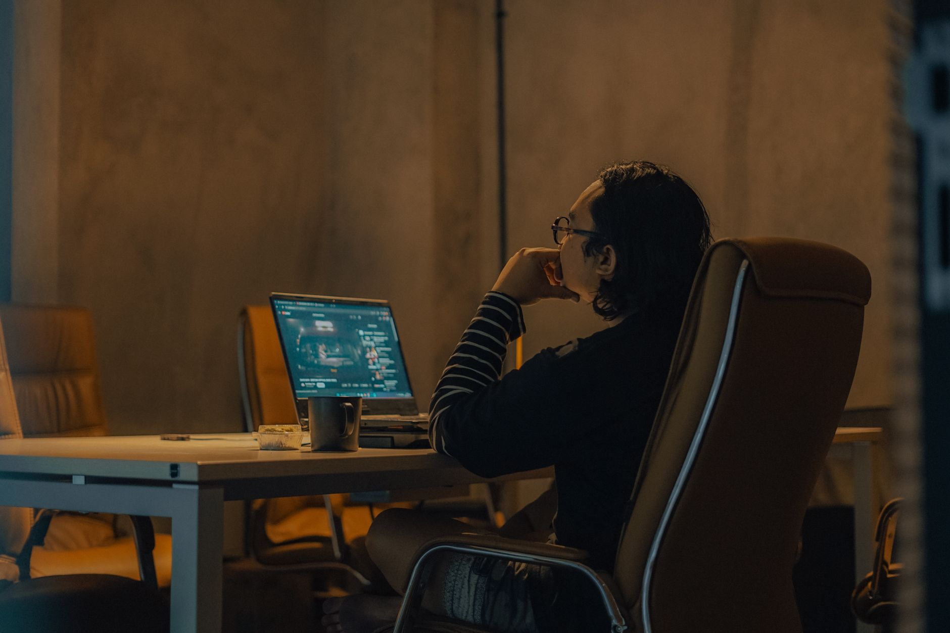 A man sits at a desk working on his laptop late in a dimly lit office, enjoying a cup of coffee.